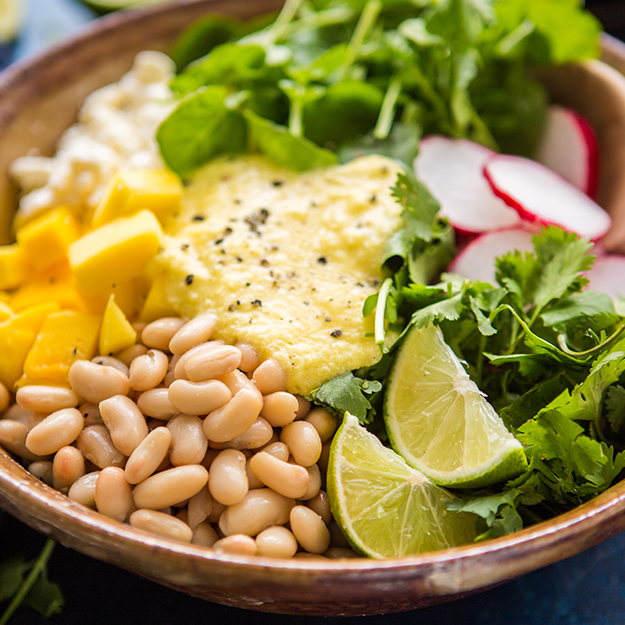Healthy bowl with white beans, mango, feta, radishes, greens, salad dressing and lime on a colorful background.