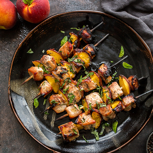 Pork and nectarine skewers on a dark plate, garnished with herbs, dark moody setting.