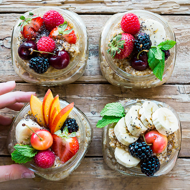 A hand holding one of four bowls of oatmeal topped with a variety of fresh fruits on a rustic wooden table.