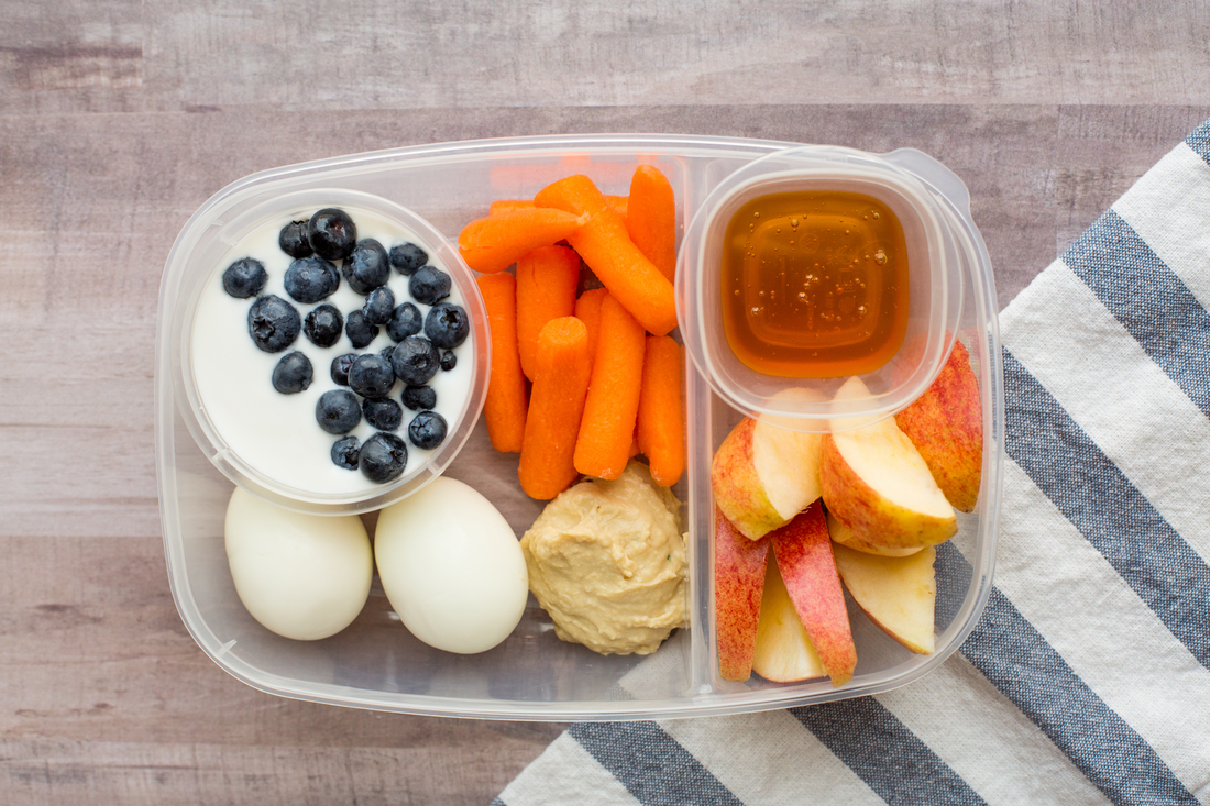 A healthy snack box with eggs, yogurt, carrots, hummus, apple, and honey on a striped napkin.