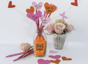 A bottle of honey with heart-shaped notes on sticks and a bouquet of roses nearby, romantic setting with pink accents.