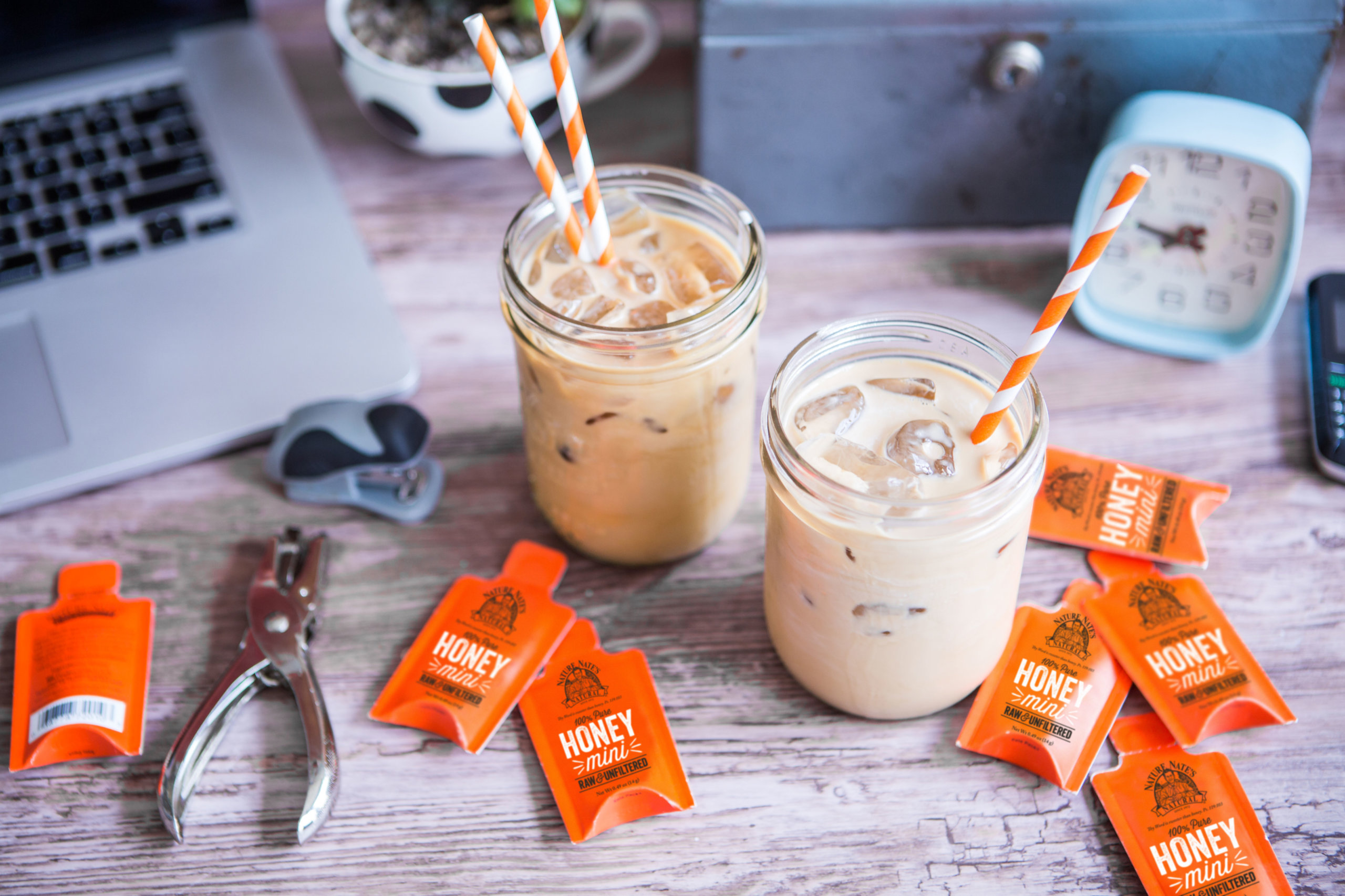 Two iced lattes with orange striped straws, Nate's honey minis scattered on a desk with laptop, clock, and keys.