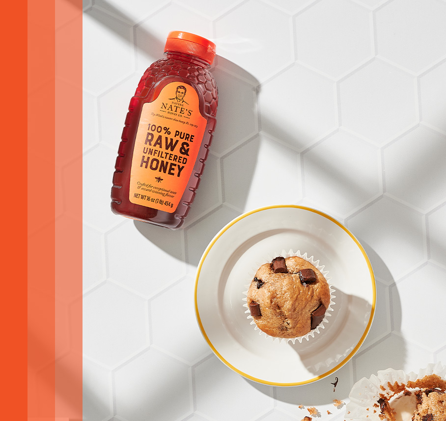 Nate's honey bottle beside a chocolate chip muffin on white hexagonal tiles.