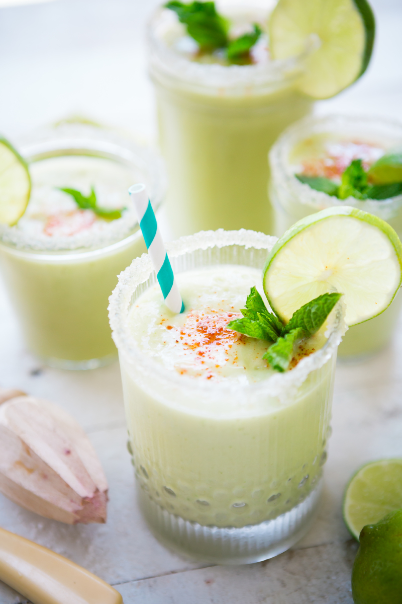 Avocado smoothies garnished with lime, mint, and a dash of spice, with straws, on a marble counter.