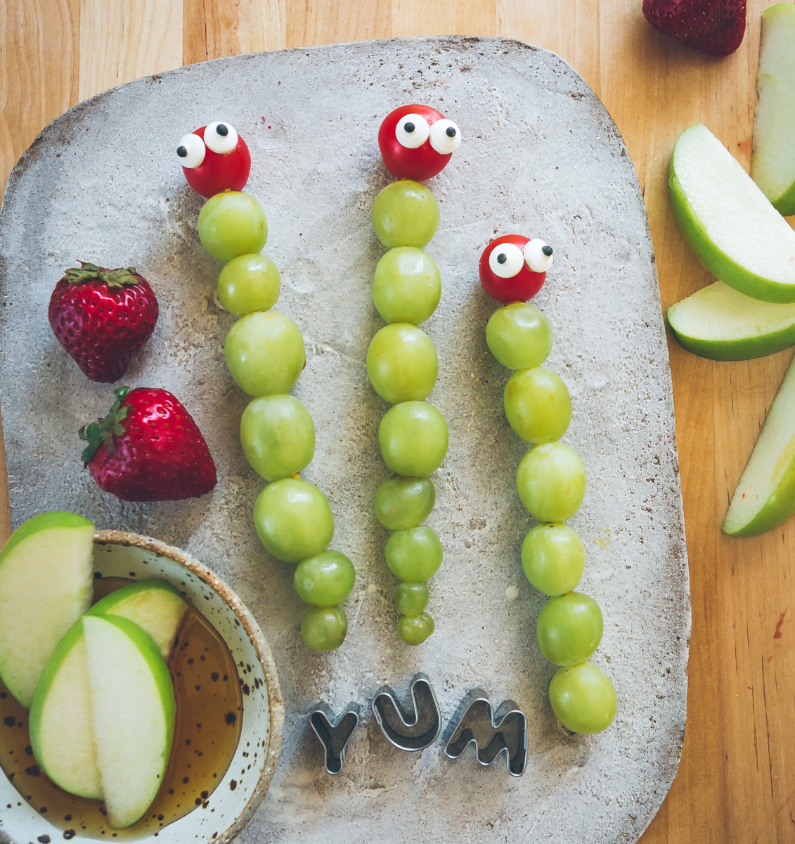 Food art on a cutting board spelling YUM, with grapes as caterpillars, cereal and apples with honey, on a wood surface.
