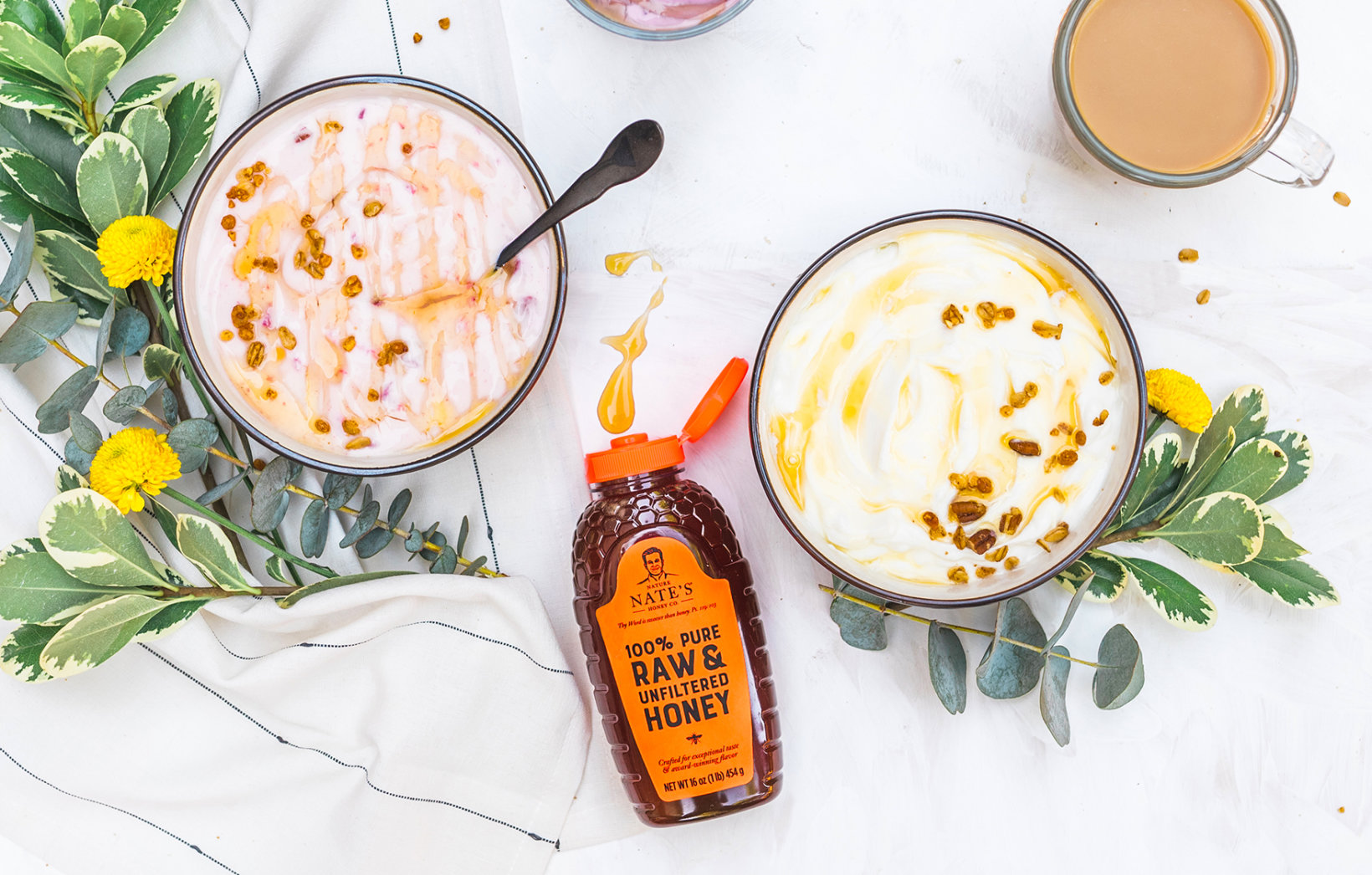 Two bowls of yogurt with drizzled honey and a bottle of Nate’s raw honey, surrounded by greenery and flowers.
