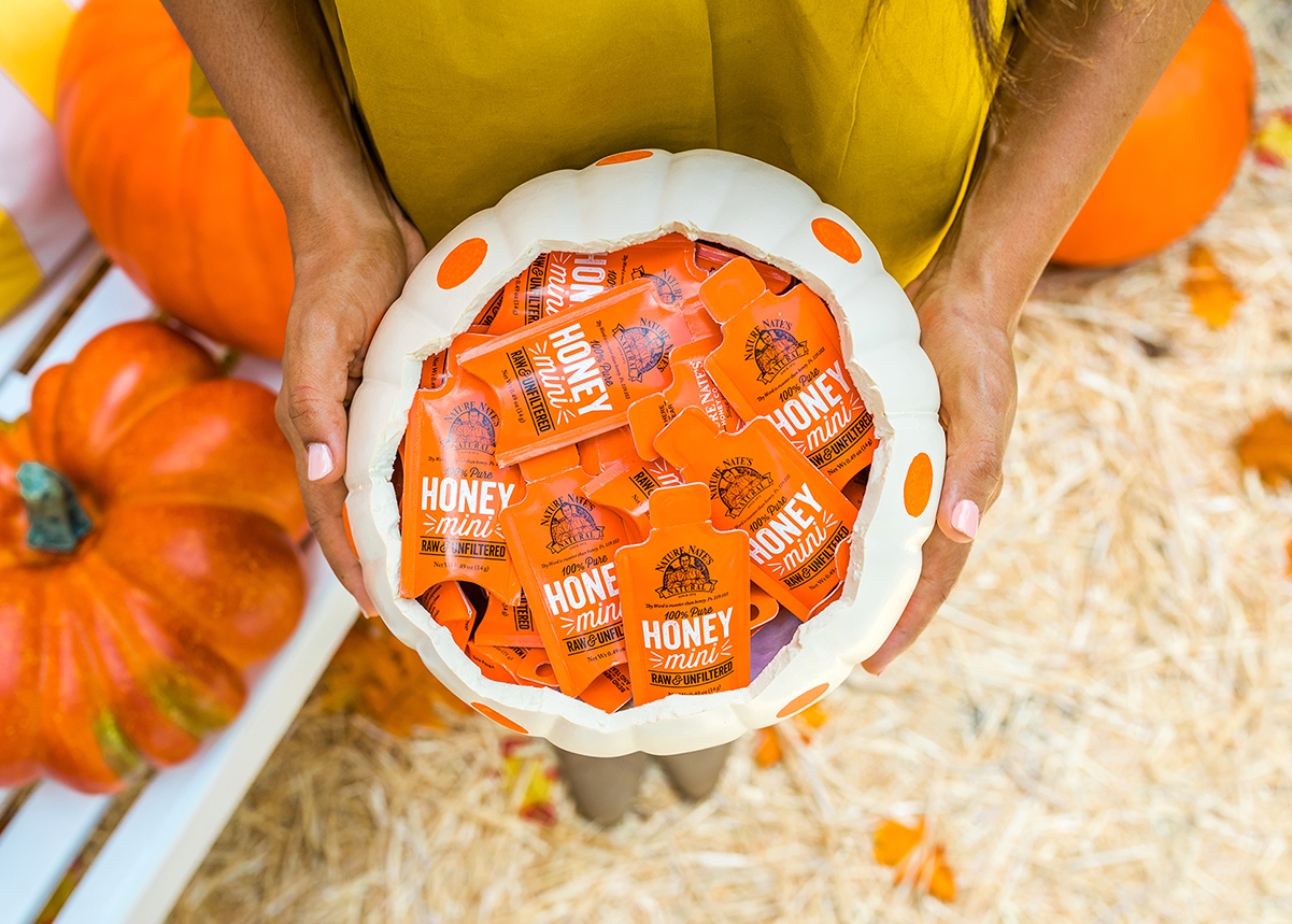 A person holding a white pumpkin basket filled with Nate's orange honey minis, surrounded by pumpkins.