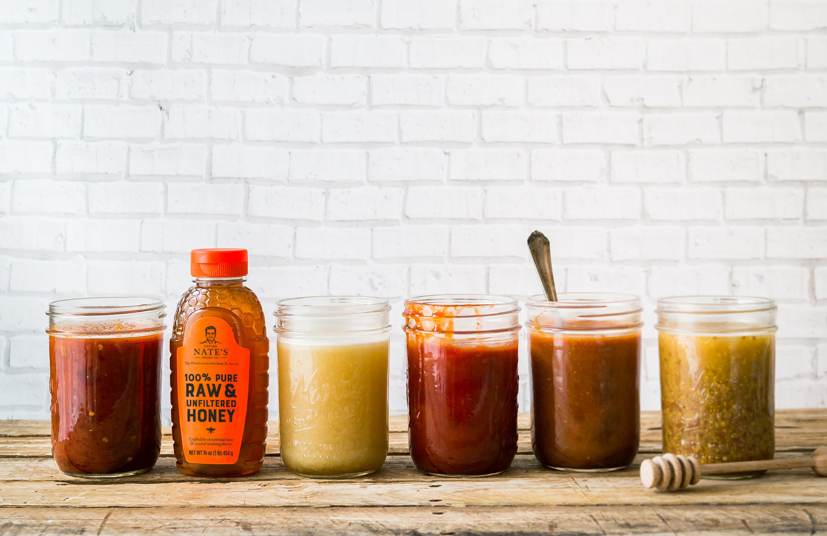 A row of assorted homemade sauces in jars with a bottle of Nate's honey, against a white brick wall.