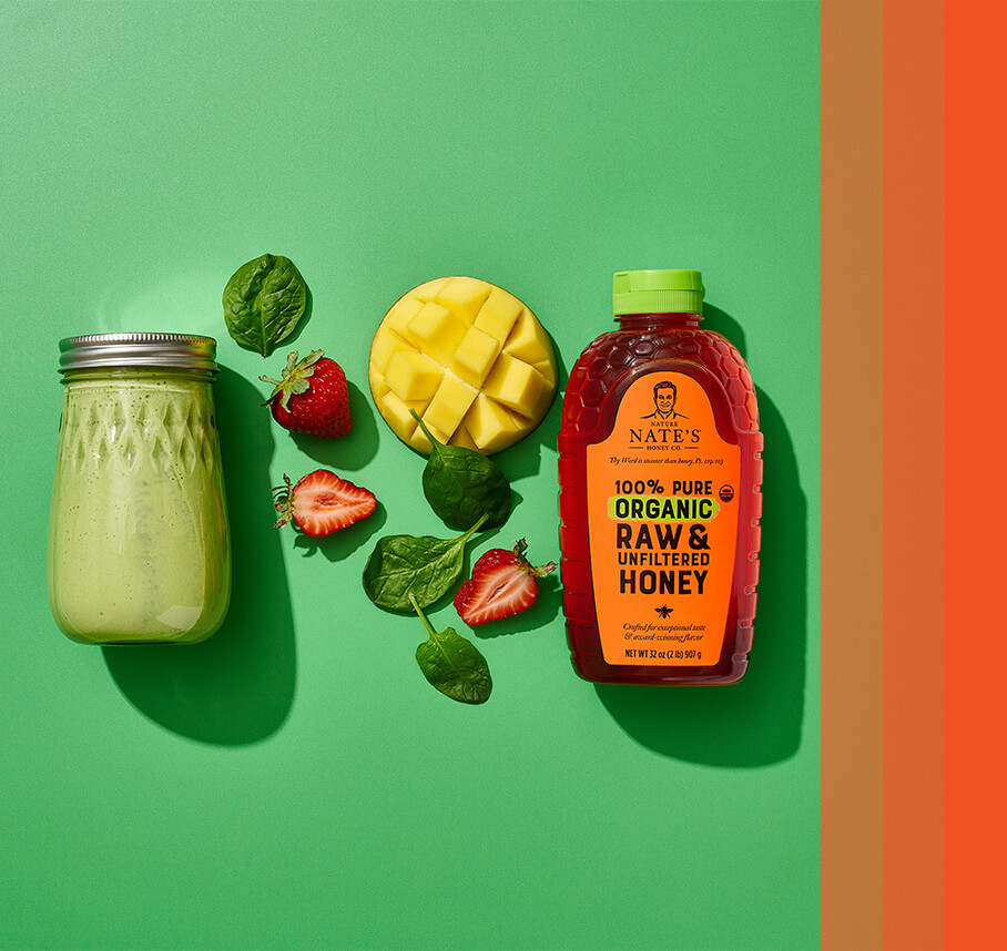 Mason jar filled with green liquid next to spinach leaves, strawberries, mango and Nate's organic honey bottle.