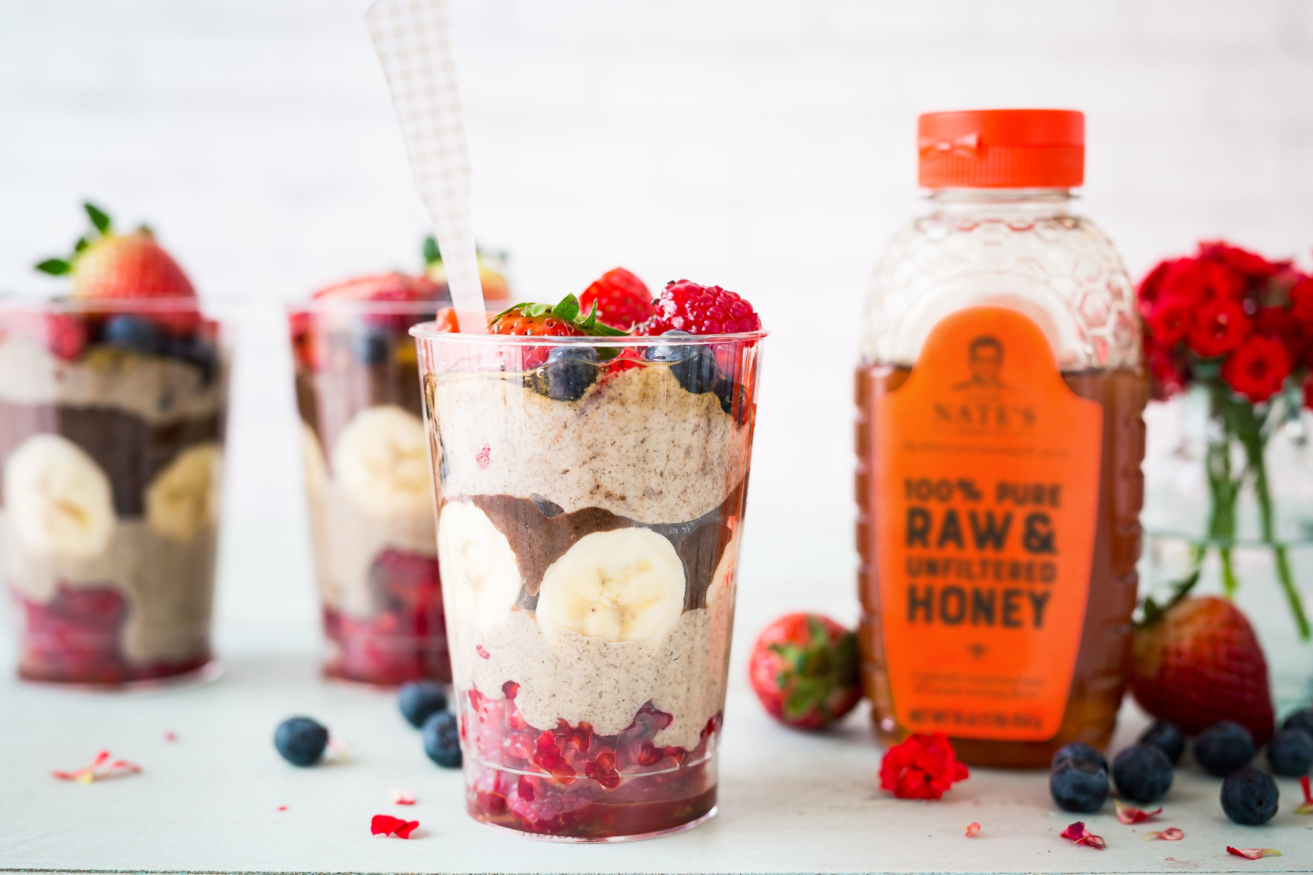 Parfaits in clear glasses with berries and bananas, with a bottle of Nate's honey and flowers in the background.