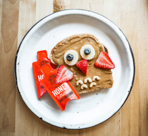 A fun owl toast with nut spread, banana and blueberry eyes, strawberry wings, next to a Nate's honey mini.