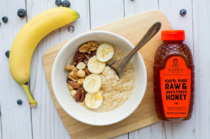 Oatmeal with sliced banana and nuts, and a bottle of Nate's honey on a wooden board.