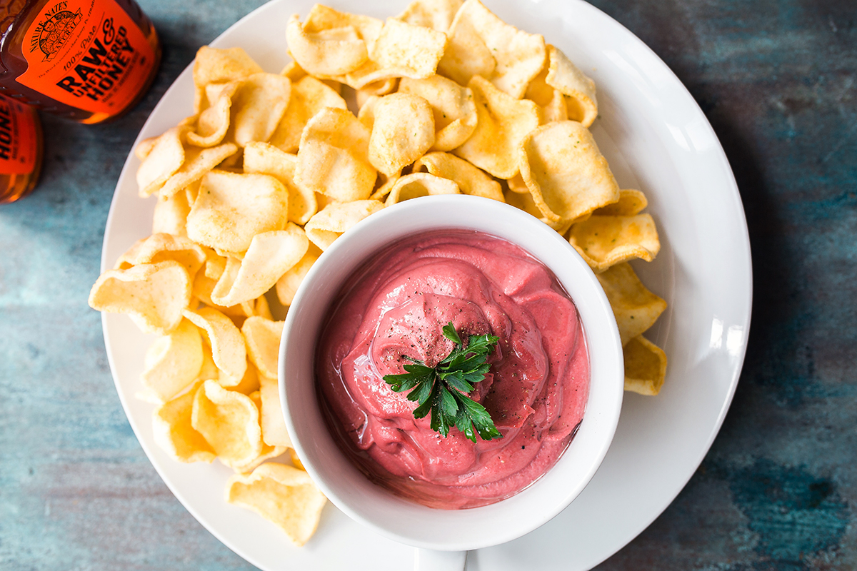 Beet dip in a bowl with chips and a bottle of Nate’s honey on a blue textured surface.