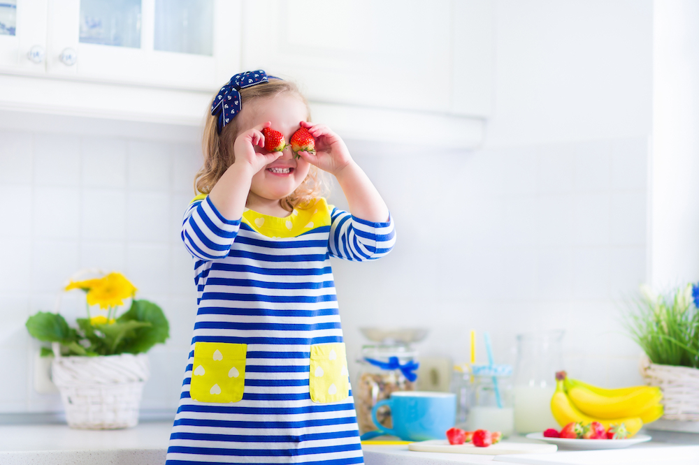 Cheerful child holding strawberries over eyes, with a kitchen setting and fruit in the background.