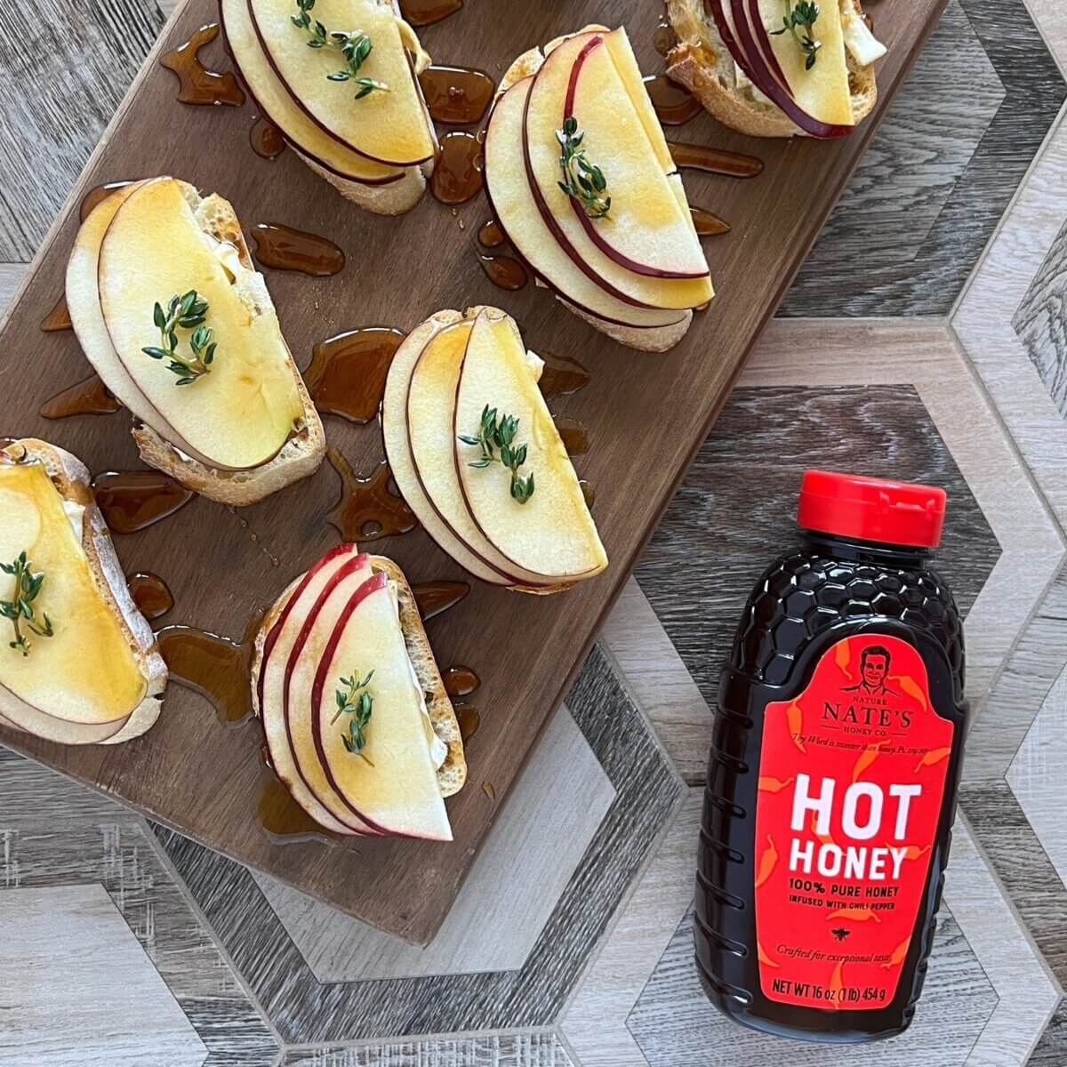 Apple brie crostini bites topped with thyme and honey on a wooden board next to a bottle of Nate’s hot honey.