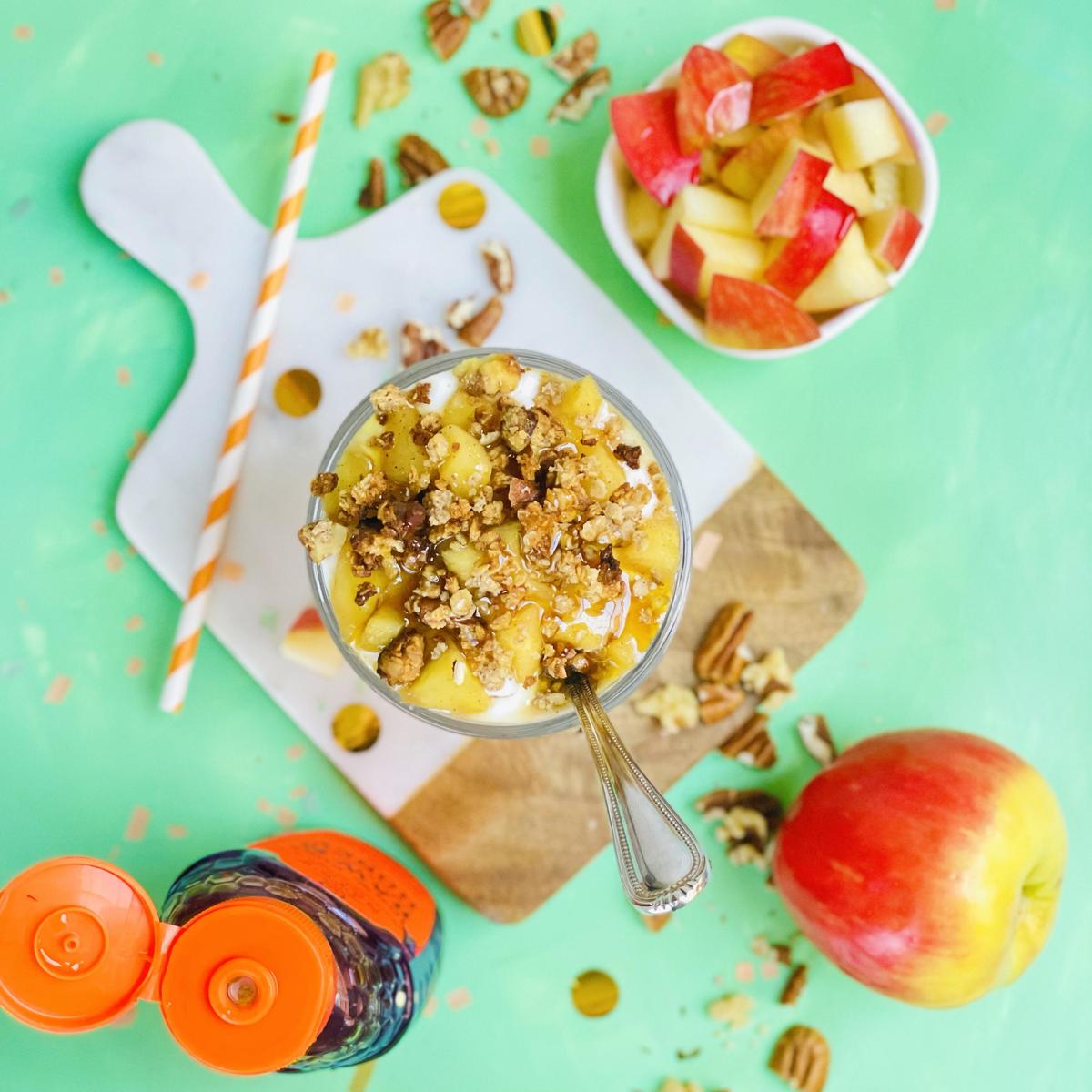 Apple parfait topped with nuts and honey, fresh fruit bowl, apple, and a bottle of Nate's honey on a green background.