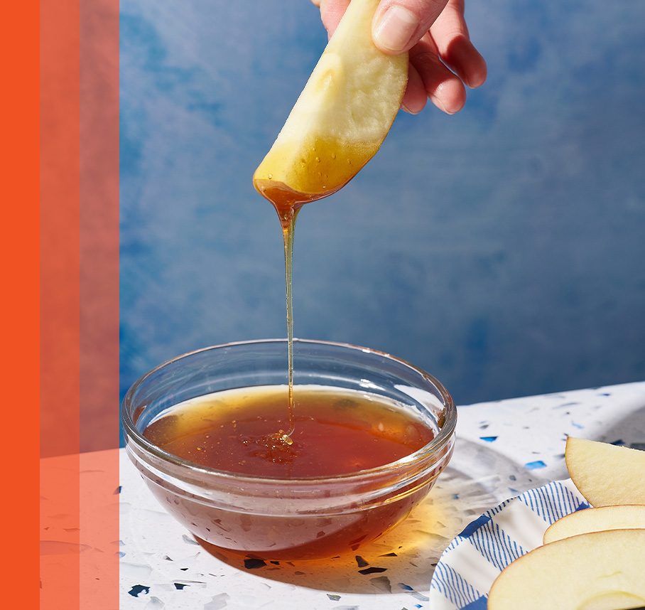 Hand dipping an apple slice into Nate’s honey in a clear glass bowl with blue background.