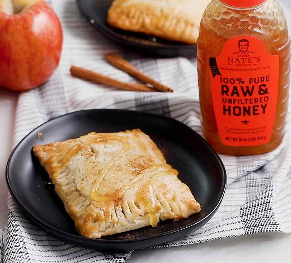 Apple turnover drizzled with honey on a plate with a bottle of Nate’s honey and cinnamon sticks on a striped cloth.