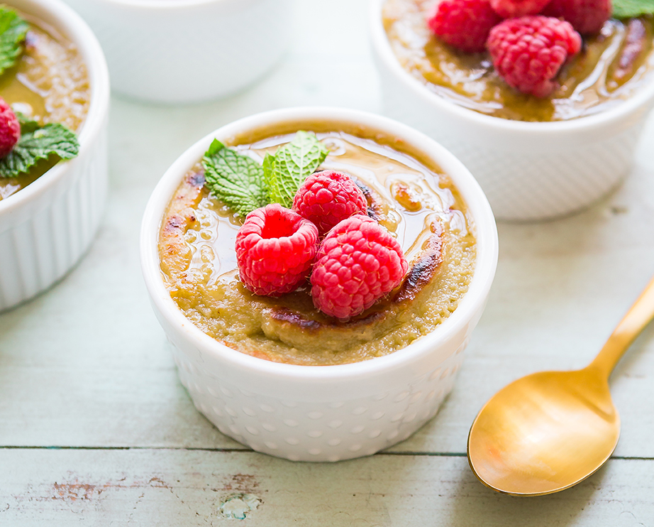Creme brulee in white ramekins garnished with raspberries with golden spoon on a pastel background.