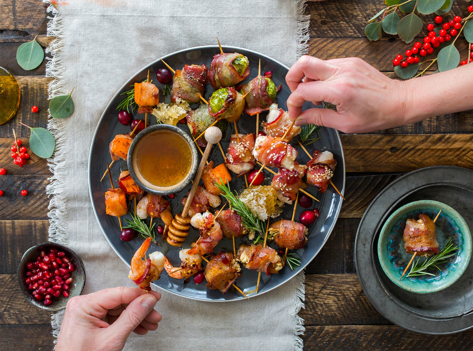 Holiday appetizer platter with skewers, dipping sauce, and hands serving, on a rustic wooden table.