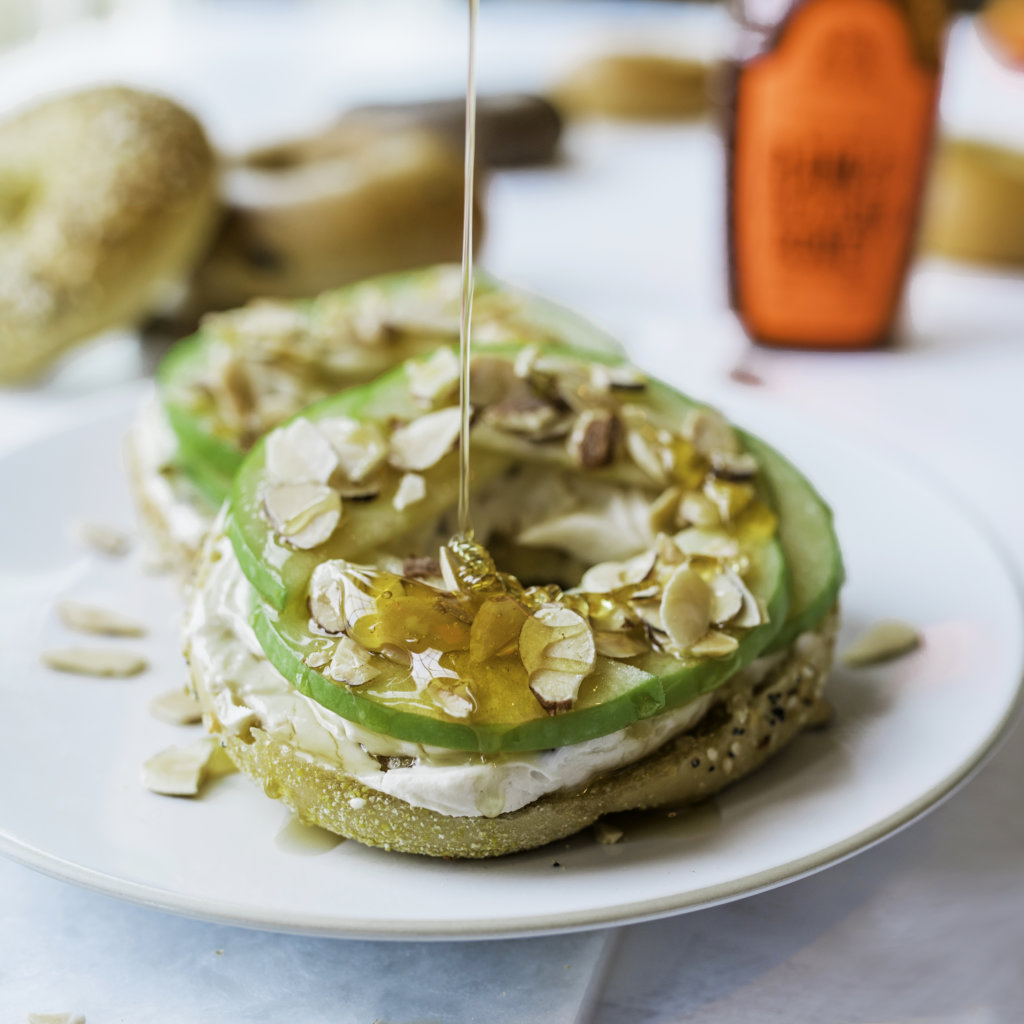 Bagel topped with green apple slices, cream cheese, slivered almonds and drizzled with honey, with Nate's honey bottle.