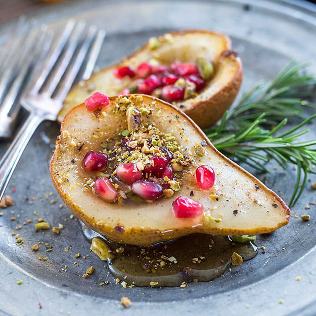 Baked pears with honey drizzle, pomegranate seeds and crushed pistachios on a pewter plate, rustic setting.