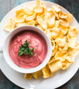 Beet dip in a bowl with chips on a blue textured surface.