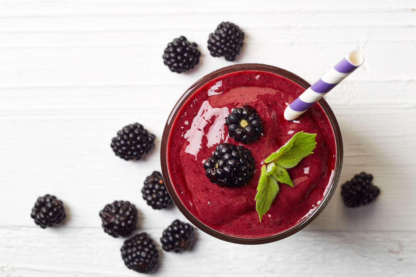 A vibrant blackberry smoothie in a glass, mint garnish, on a white wooden surface with scattered berries.