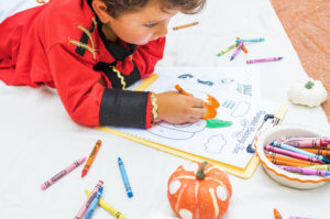 A child focused on coloring a Halloween-themed activity sheet surrounded by scattered crayons on the floor.