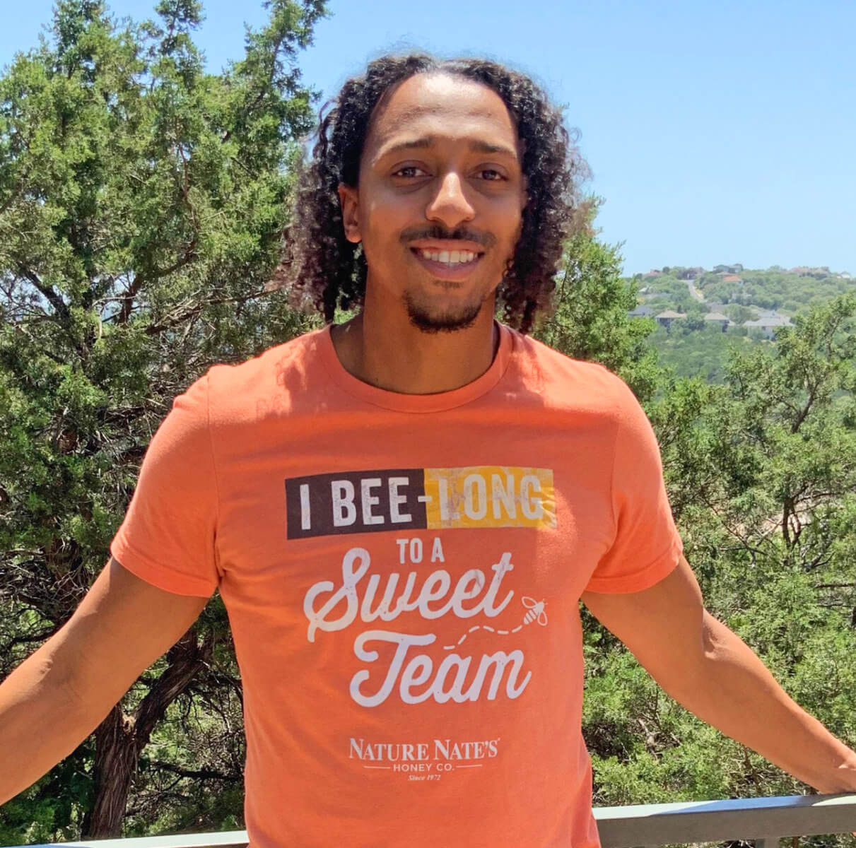 A smiling Nate's employee wearing a t-shirt with a bee and sweet team slogan, outdoors.