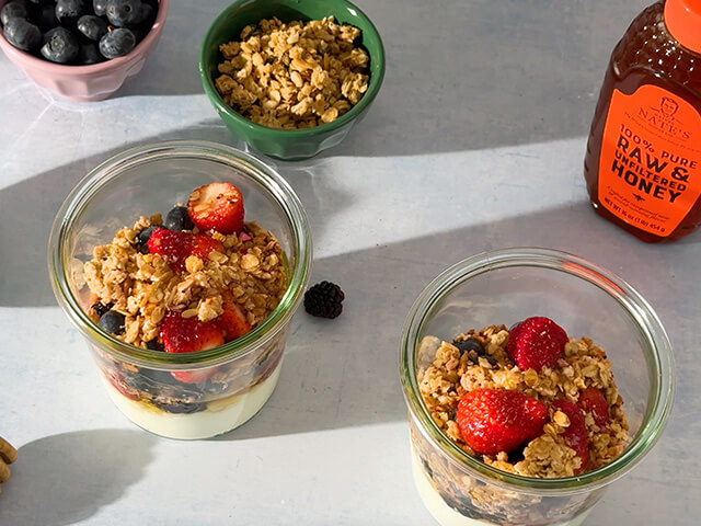 Two parfaits of yogurt, granola, berries and honey in glass cups. Next to two prep containers of granola and blueberries.