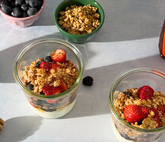 Two parfaits of yogurt, granola, berries and honey in glass cups. Next to two prep containers of granola and blueberries.