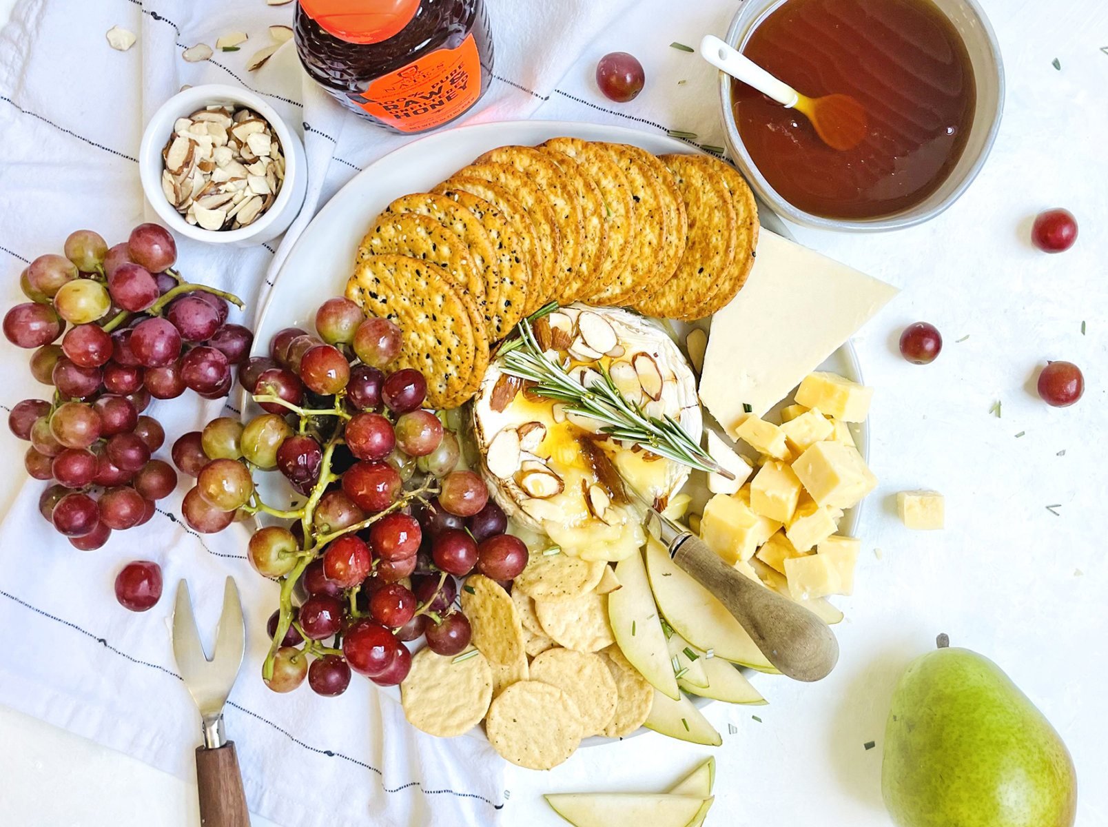 A cheese platter with grapes, crackers, nuts, Nate’s honey, and a pear on a white cloth background.