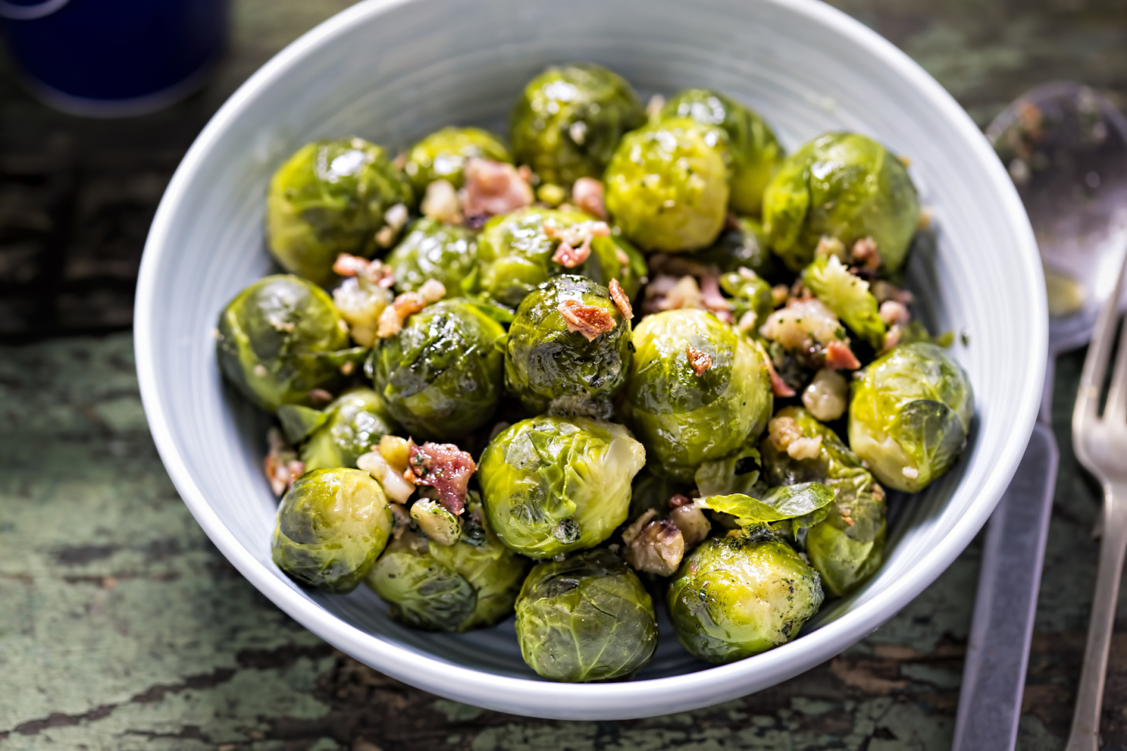 A bowl of cooked Brussels sprouts mixed with onion and bacon bits and glazed with honey, on a rustic, greenish surface.