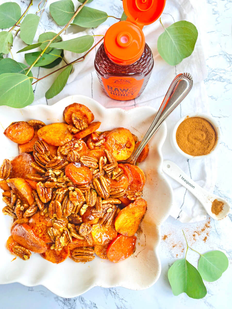 A dish of candied yams and pecans next to a Nate’s honey bottle with an orange cap, with a spoon of cinnamon.