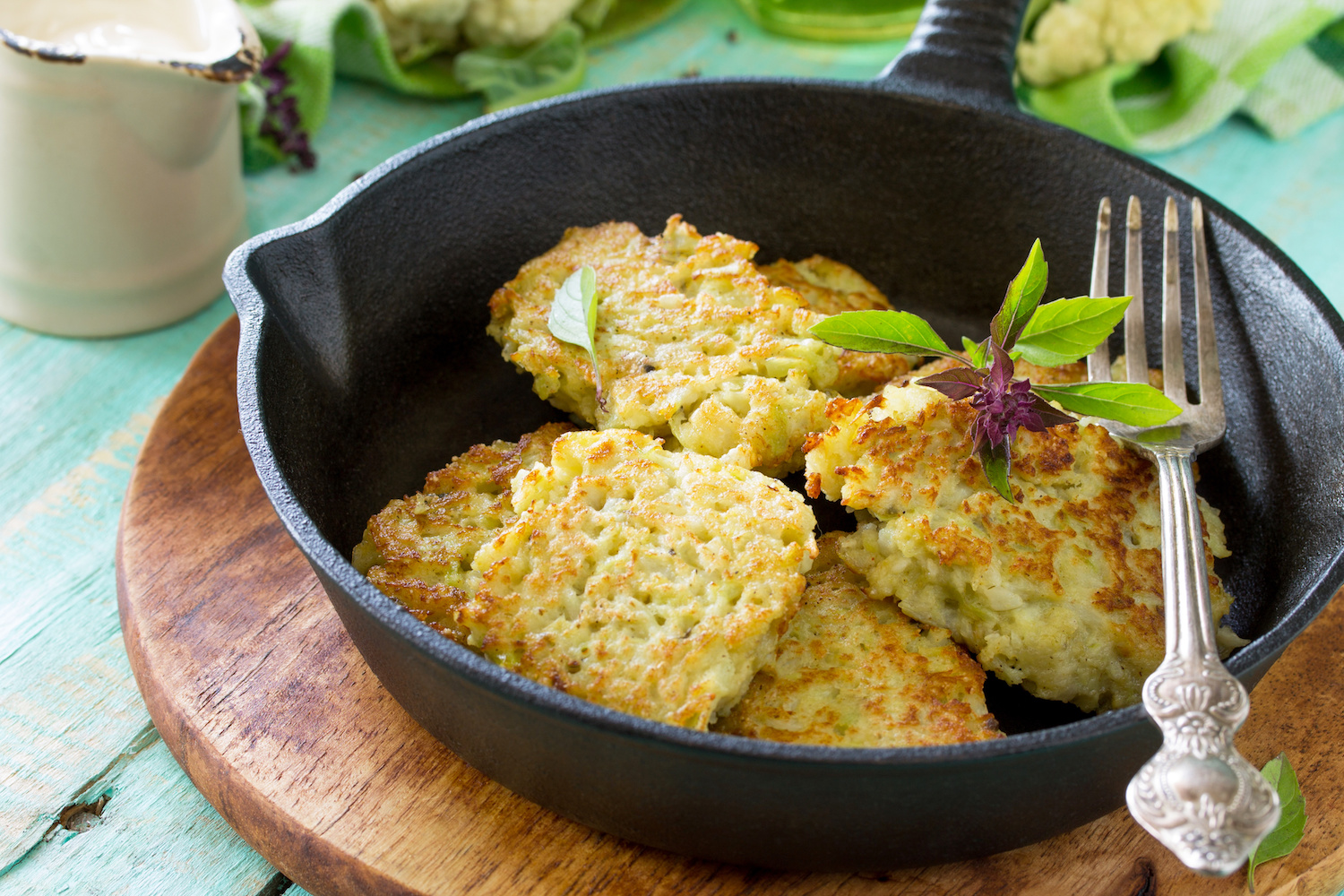A skillet with golden-brown cauliflower fritters garnished with basil, ready to be served on a rustic wooden table.