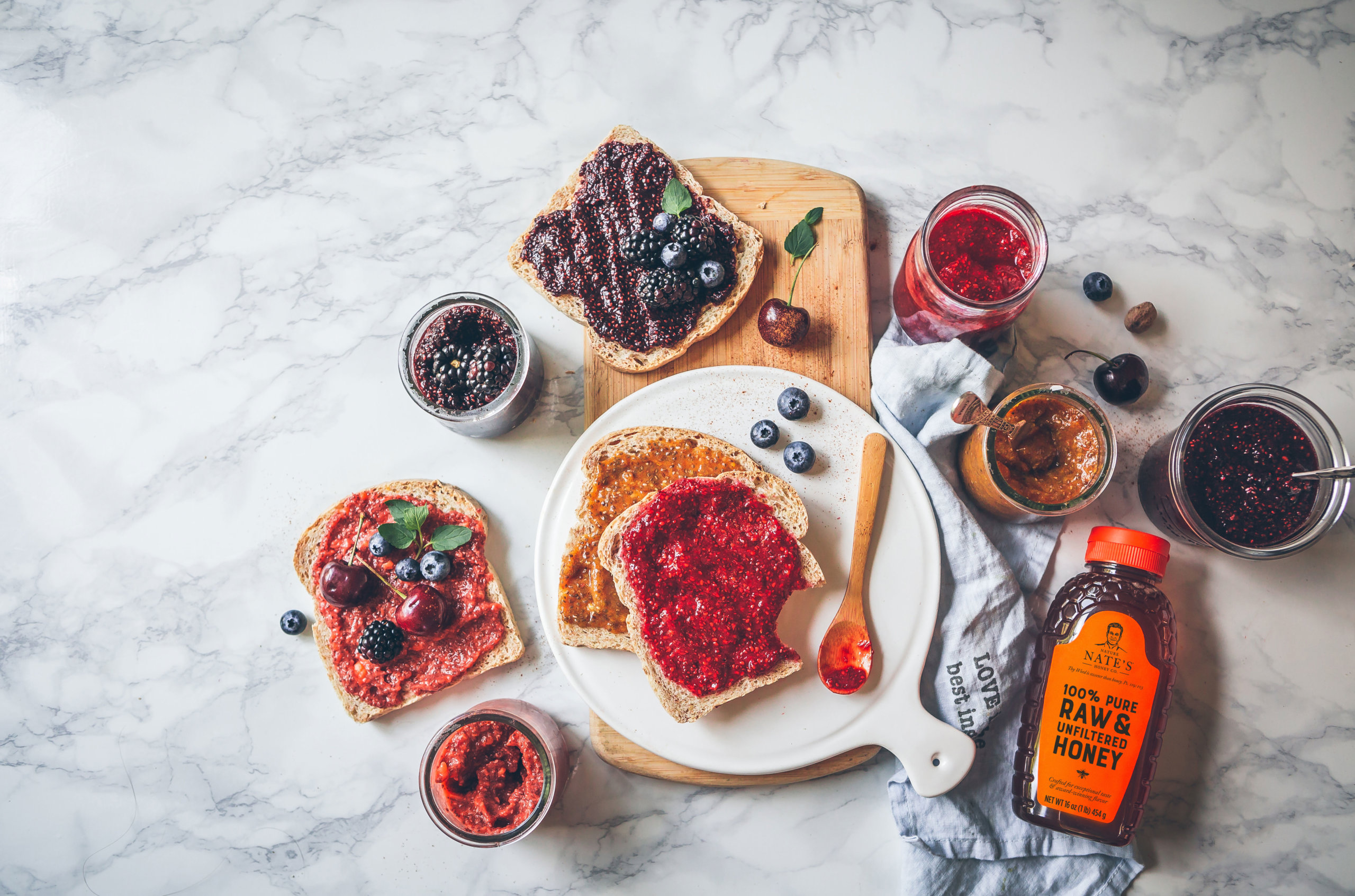 Toast topped with berry jam beside a bottle of Nate's honey and fresh fruit on a marble surface.