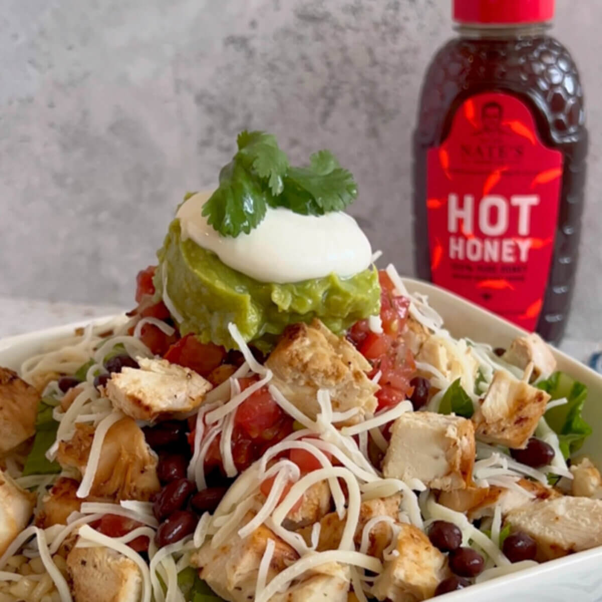 Cubes of grilled chicken in a bowl, topped with cheese, guacamole and sour cream, next to a bottle of Nate's hot honey.