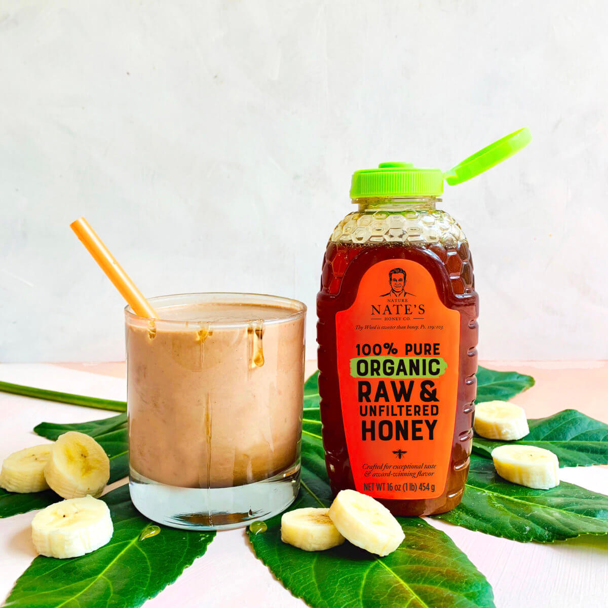 Chocolate and banana smoothie in a glass, Nate's organic honey bottle with banana slices on a leafy backdrop.