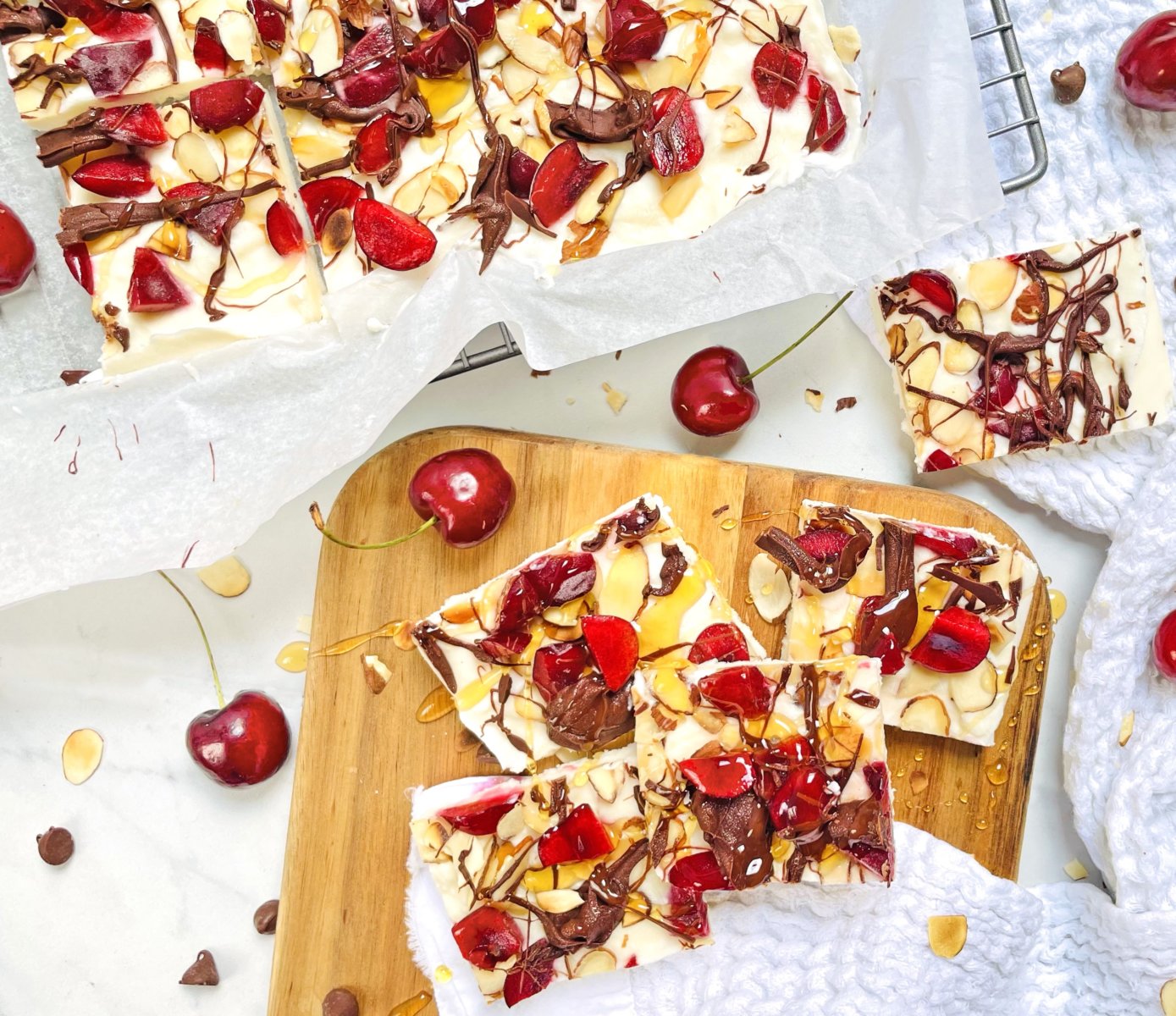 White chocolate bark with cherries and nuts, drizzled with chocolate, on a wooden board.