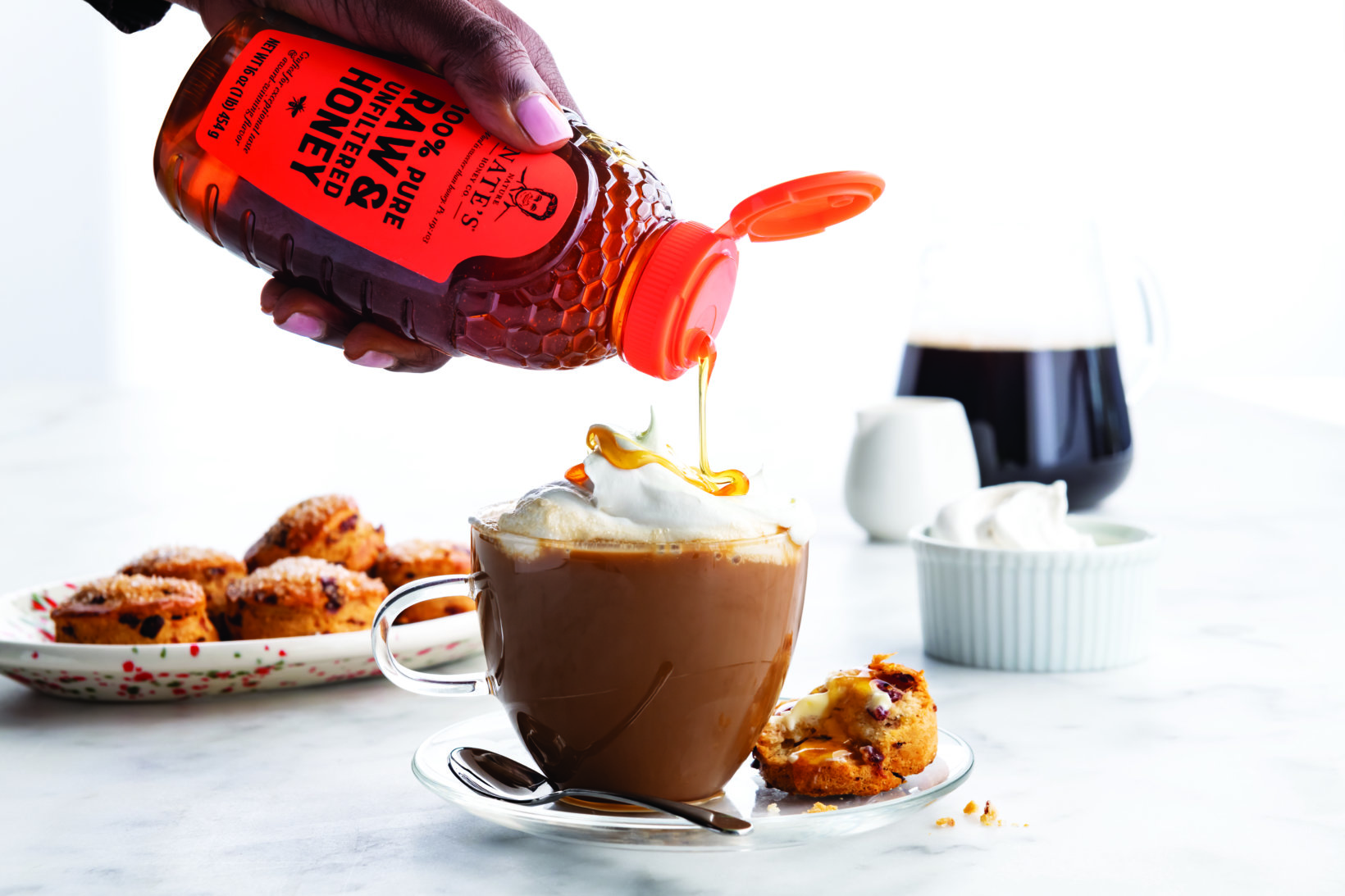 Honey from a Nate’s honey bottle being drizzled into coffee, with cookies and whipped cream in the background on marble.