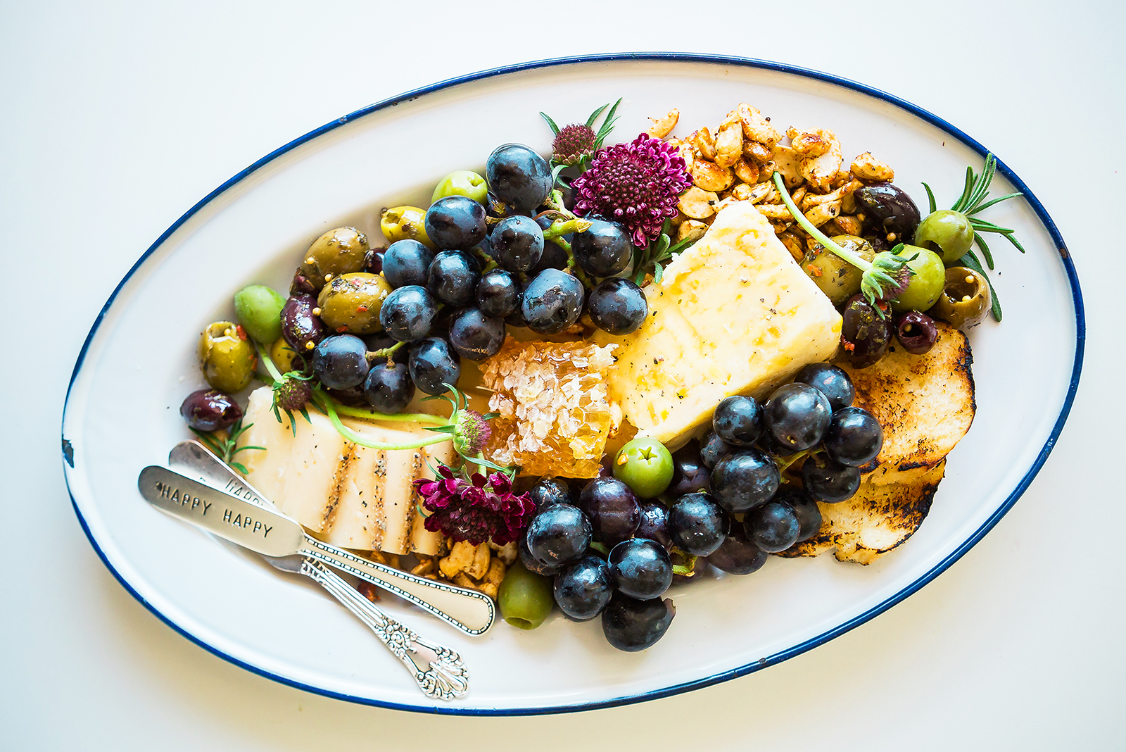 Cheese platter with grapes, olives, honeycomb, cheeses, toast and an engraved knife on a blue-rimmed plate.