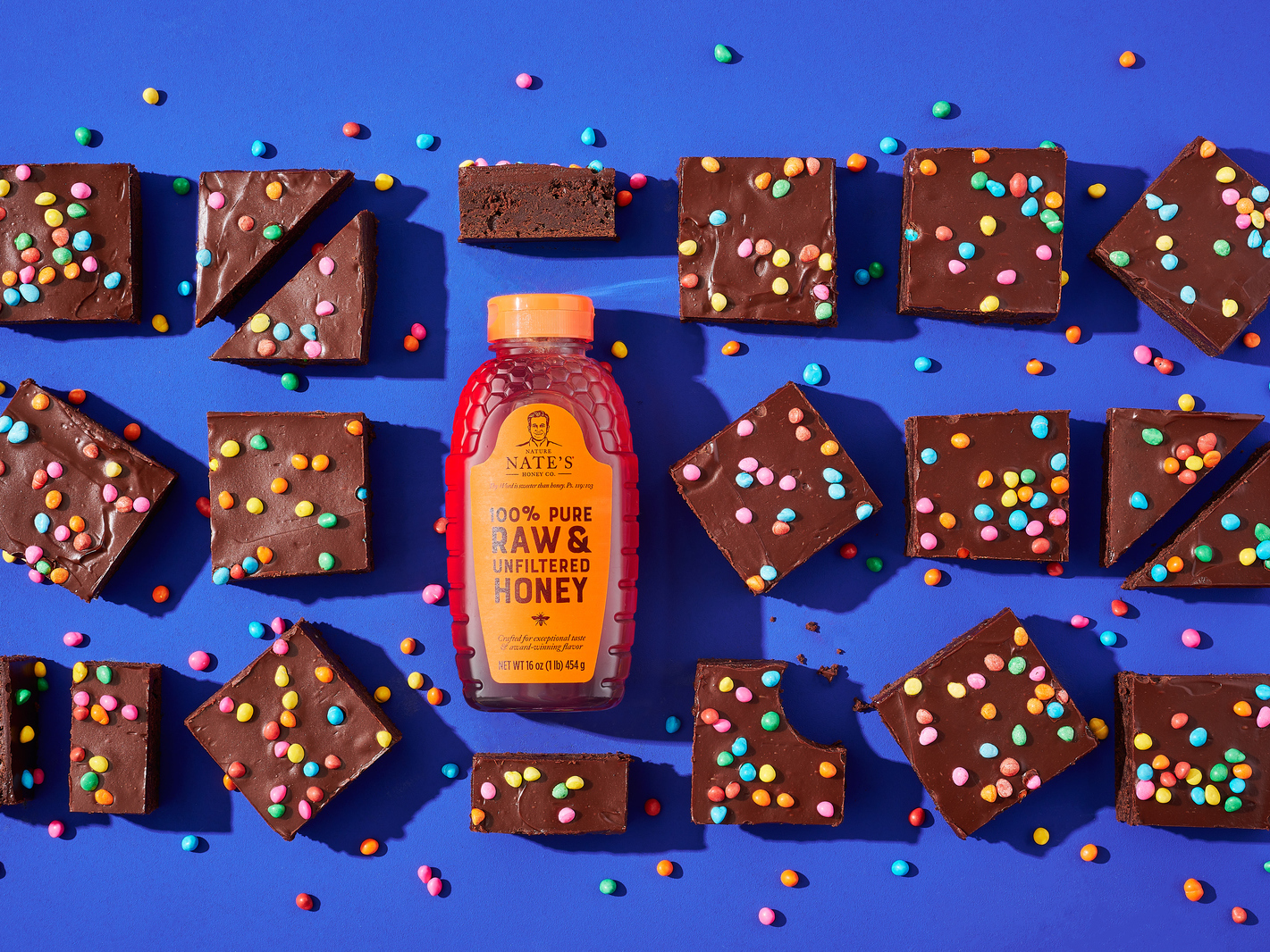 Chocolate brownies with colorful sprinkles and a Nate’s honey bottle on blue background.