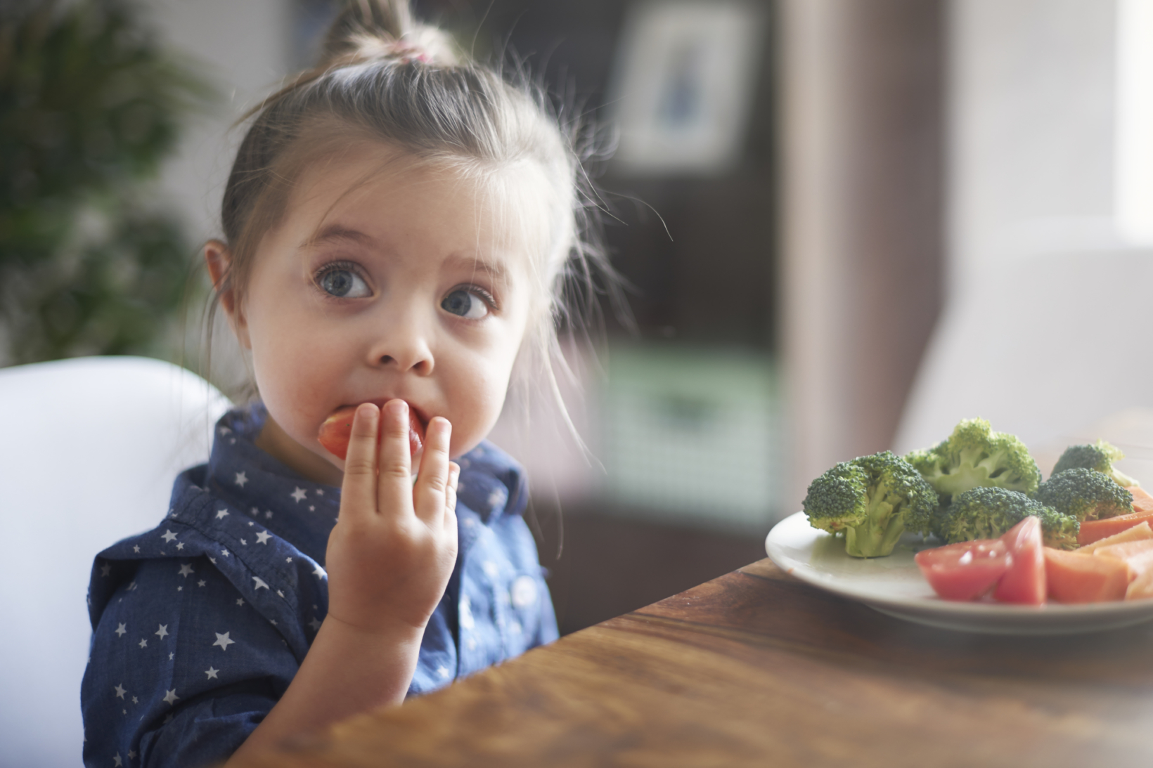 Surprised little girl at a table with a plate of veggies in a home setting.