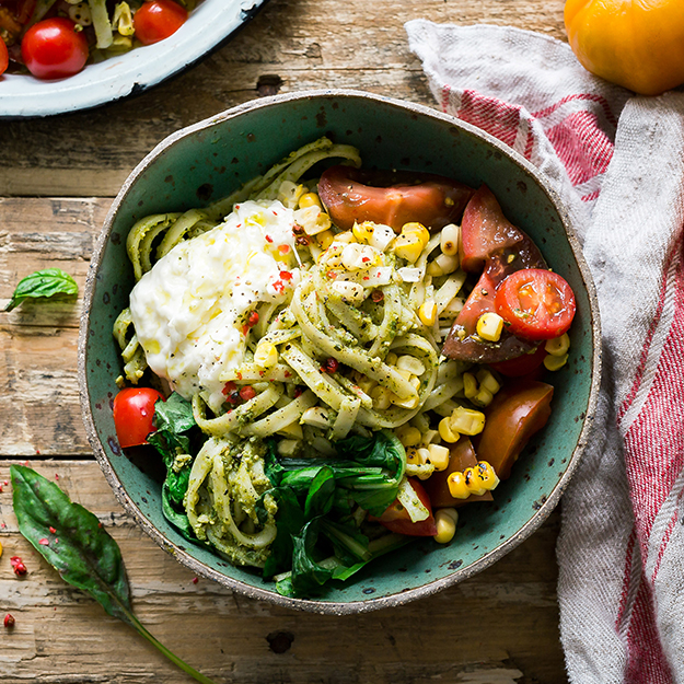 Pesto pasta with fresh veggies and burrata cheese, beside a striped napkin on a wooden table.