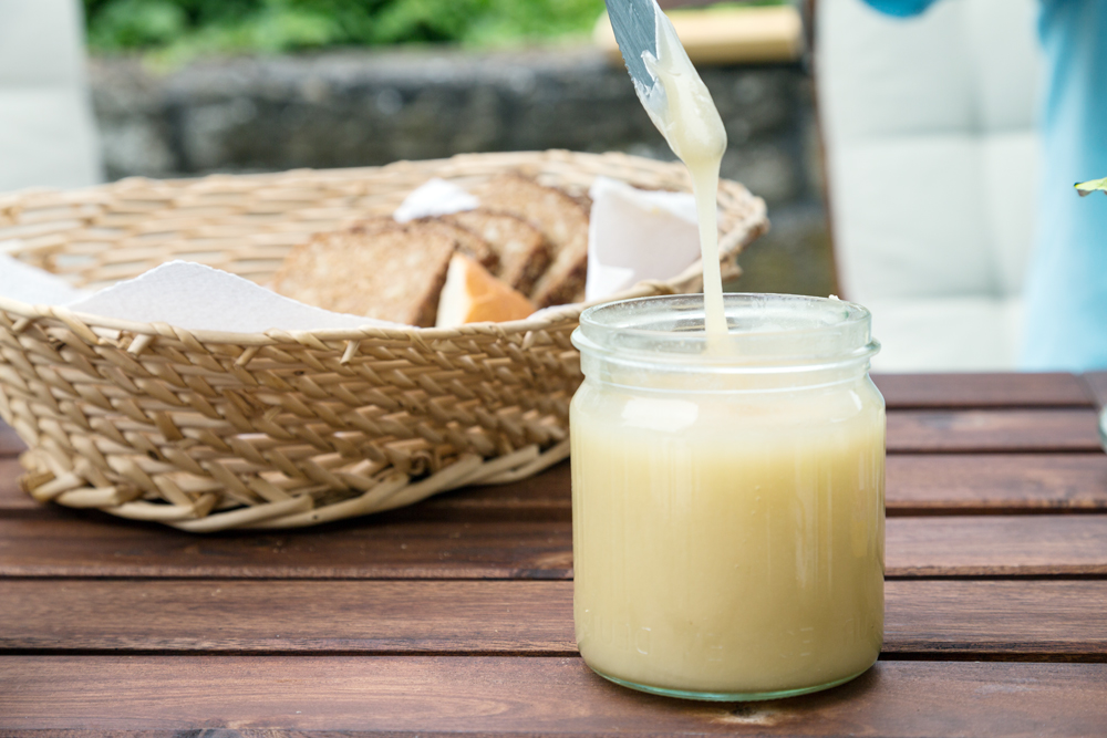 Jar of creamy honey with a drizzler and bread basket on a wooden table.