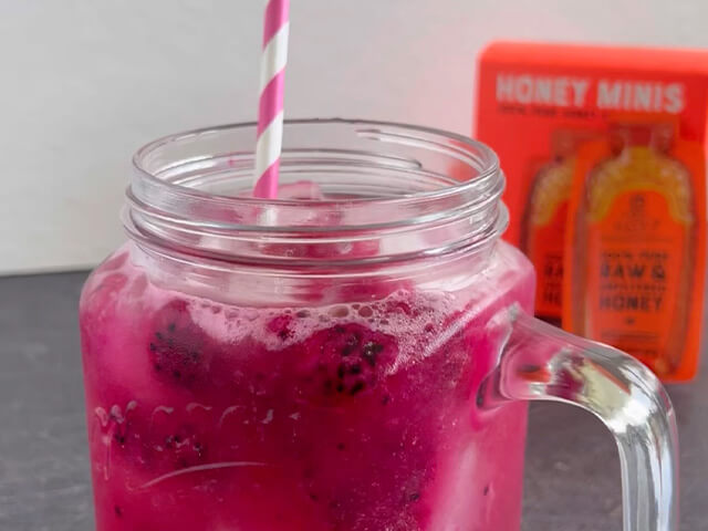 Mason jar cup containing purple dragon fruit beverage. Pink and white striped straw in cup and honey minis box in background.
