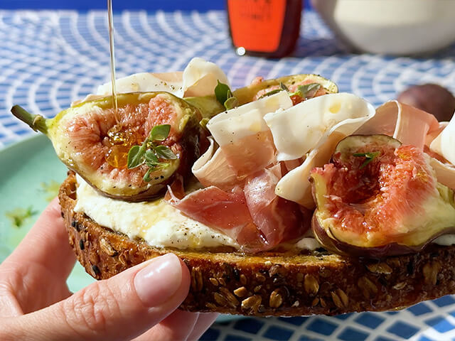 Hand holding toast loaded with toppings. Toppings are fig, prosciutto, burrata and honey drizzle.