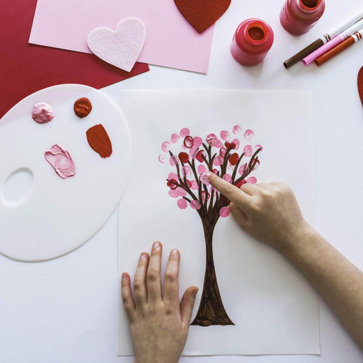 Finger painting a tree with fingerprint leaves on paper, palette and pink paint on a white and red background.