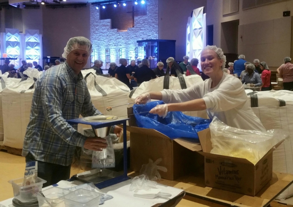 Two smiling people wearing hairnets and casual clothing are packaging food at a table, with large bags.